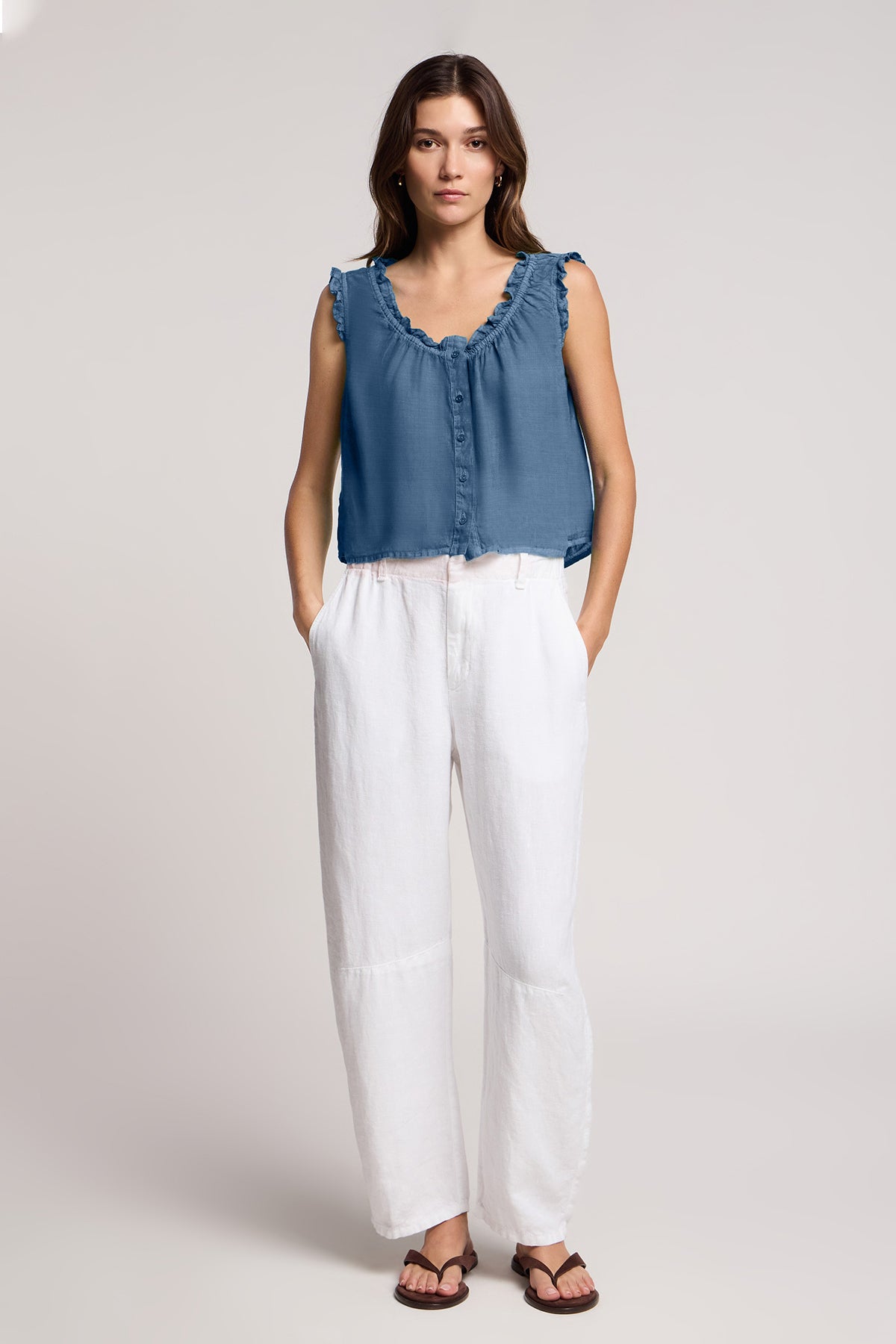A woman stands against a plain background wearing the TANA LINEN TOP by Velvet by Graham & Spencer—a feminine, sleeveless blue linen top—paired with white linen pants and sandals, her hands tucked in her pockets.