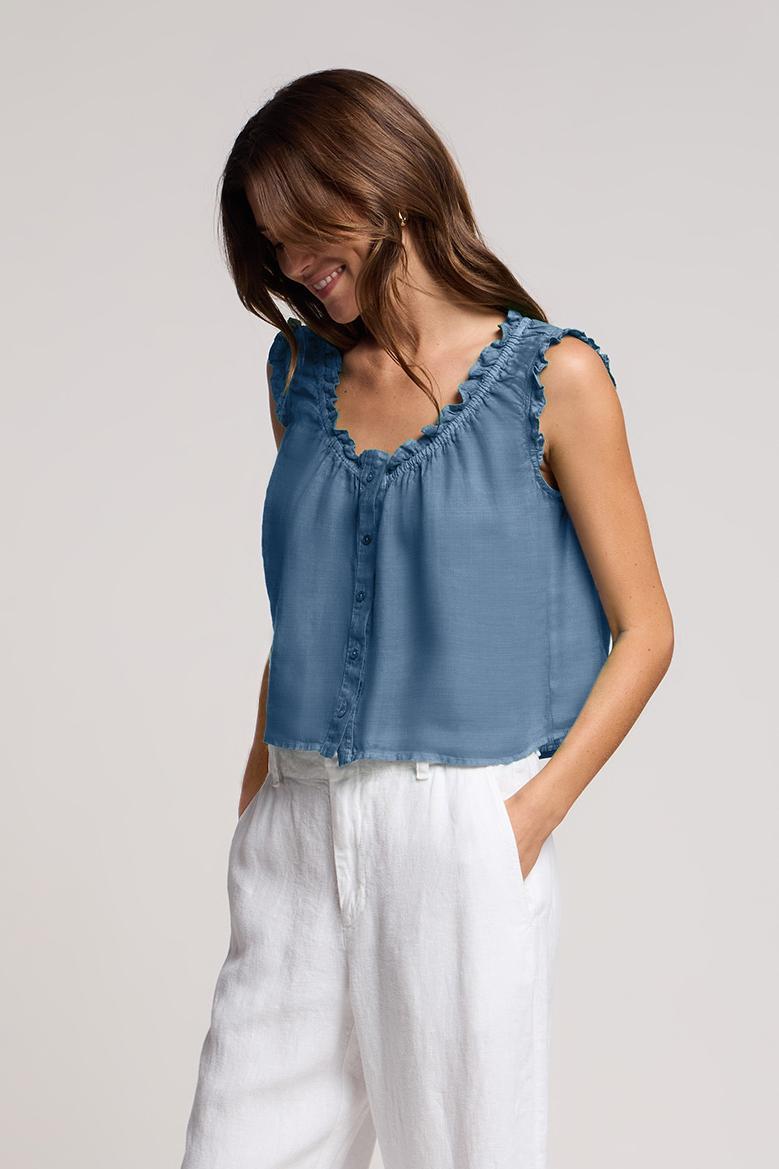 A woman wearing the TANA LINEN TOP by Velvet by Graham & Spencer— a sleeveless, button-front linen top with delicate ruffle trim—paired with white high-waisted pants, stands against a neutral background.-42118583091393
