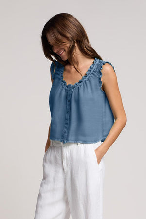 A woman wearing the TANA LINEN TOP by Velvet by Graham & Spencer— a sleeveless, button-front linen top with delicate ruffle trim—paired with white high-waisted pants, stands against a neutral background.