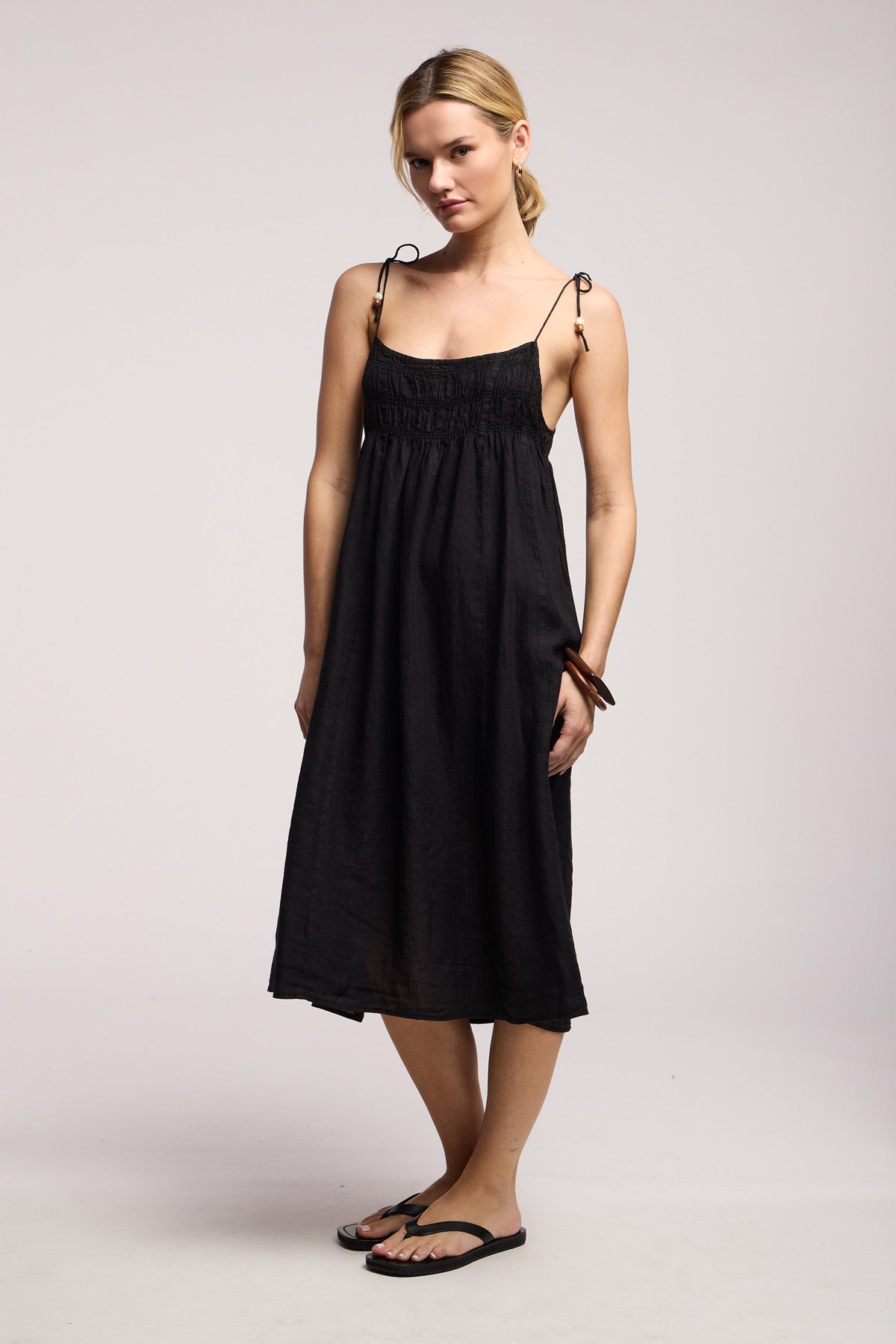 A woman stands against a plain background wearing the Velvet by Graham & Spencer TERESA LINEN DRESS, paired with black sandals and a brown bracelet.-41645183402177