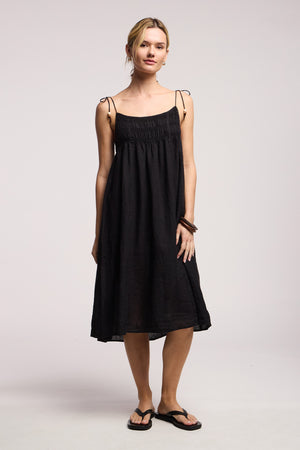 A woman with light hair pulled back and a neutral expression wears the TERESA LINEN DRESS by Velvet by Graham & Spencer, featuring tie straps, along with black flip-flops and a brown bracelet. She stands facing forward.