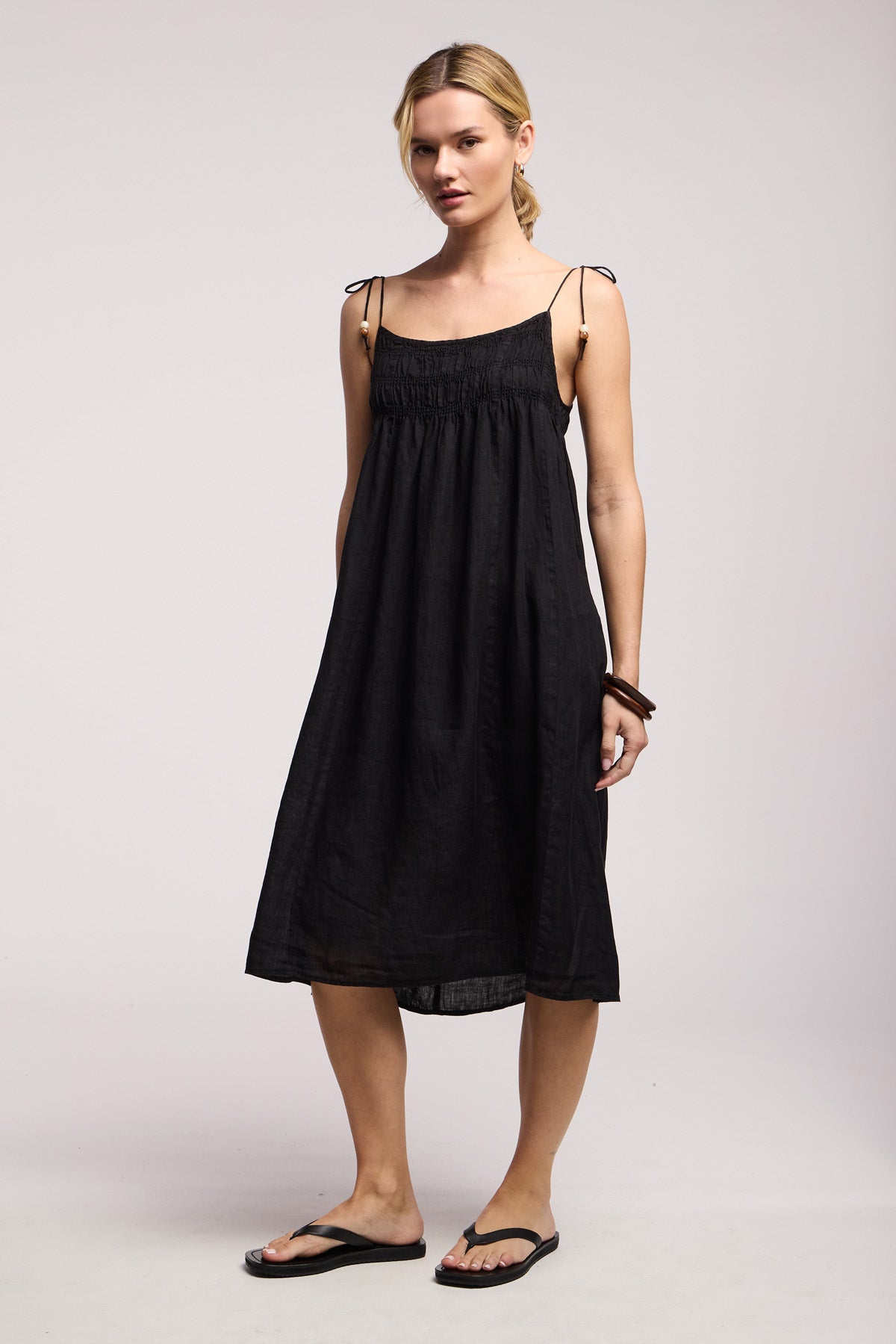 A woman stands against a plain background wearing the Velvet by Graham & Spencer TERESA LINEN DRESS, featuring black woven linen, a smocked elastic bust, tie straps, and black flip-flops.-41645183467713