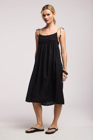 A woman stands against a plain background wearing the Velvet by Graham & Spencer TERESA LINEN DRESS, featuring black woven linen, a smocked elastic bust, tie straps, and black flip-flops.