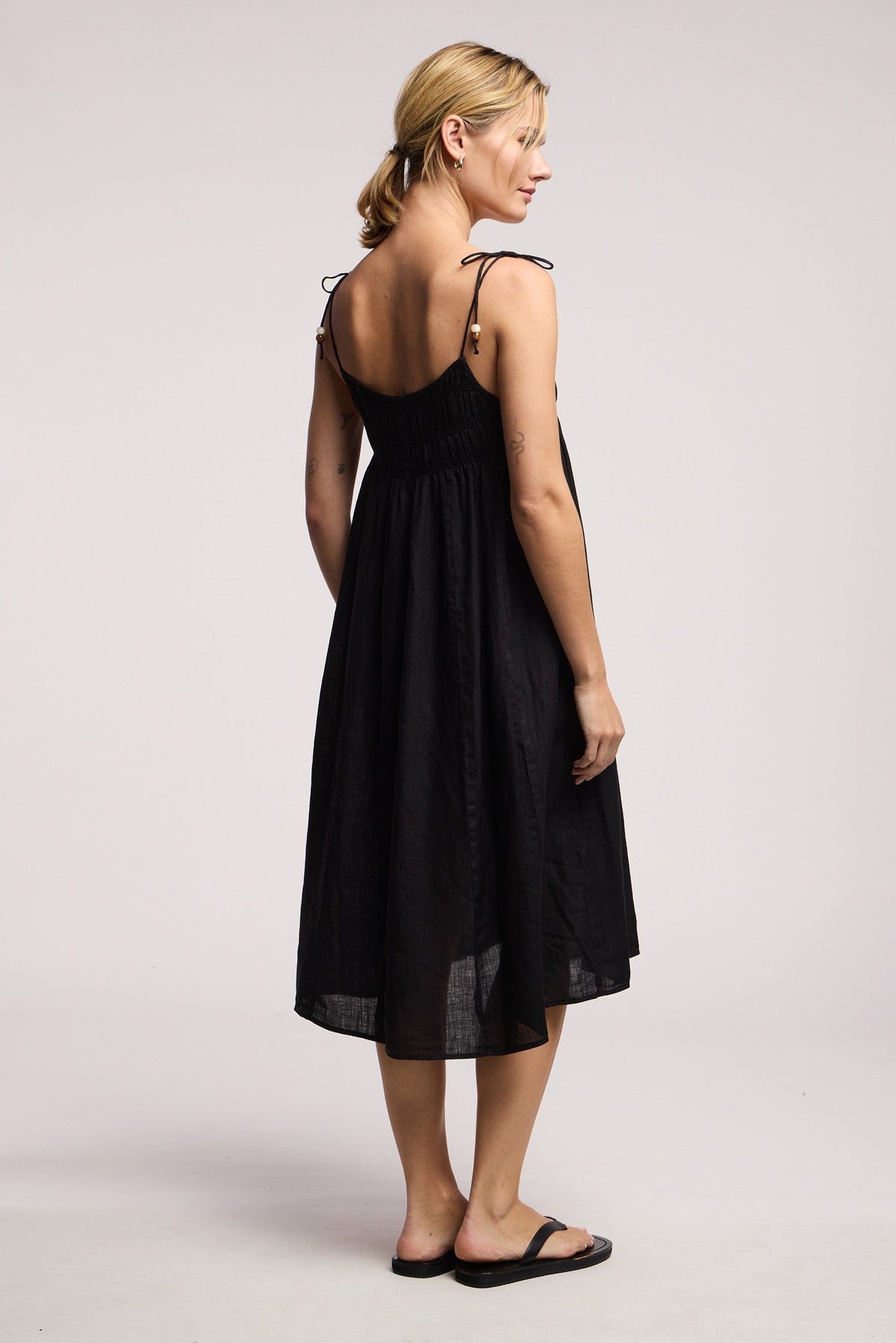 A woman stands facing away in the Velvet by Graham & Spencer TERESA LINEN DRESS—a woven linen midi featuring tie straps, a smocked elastic bust, and black sandals—set against a plain light background.-41645183566017