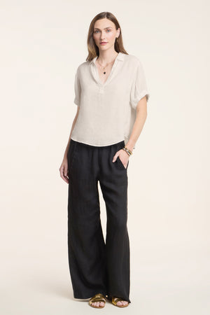 A woman stands against a light background wearing the VIVICA LINEN TOP by Velvet by Graham & Spencer—a loose beige short-sleeve lightweight linen top with a split neckline—paired with black wide-leg pants, gold sandals, and layered jewelry for an effortless warm-weather look.