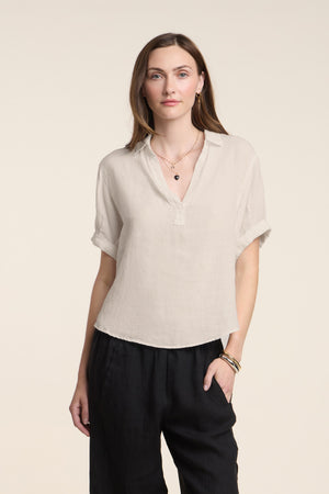 A woman wears the VIVICA LINEN TOP by Velvet by Graham & Spencer—a light beige, lightweight linen piece featuring a split neckline—paired with black pants, standing against a plain off-white background with a neutral expression; an ideal choice for effortless warm-weather dressing.