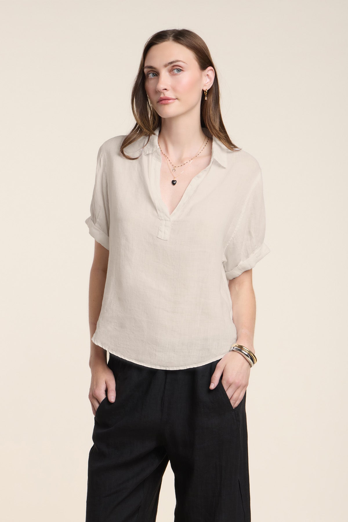 A woman stands against a plain background, wearing the VIVICA LINEN TOP by Velvet by Graham & Spencer—a light beige, split neckline short-sleeve blouse—paired with black pants and her hands in her pockets for an effortless warm-weather look.