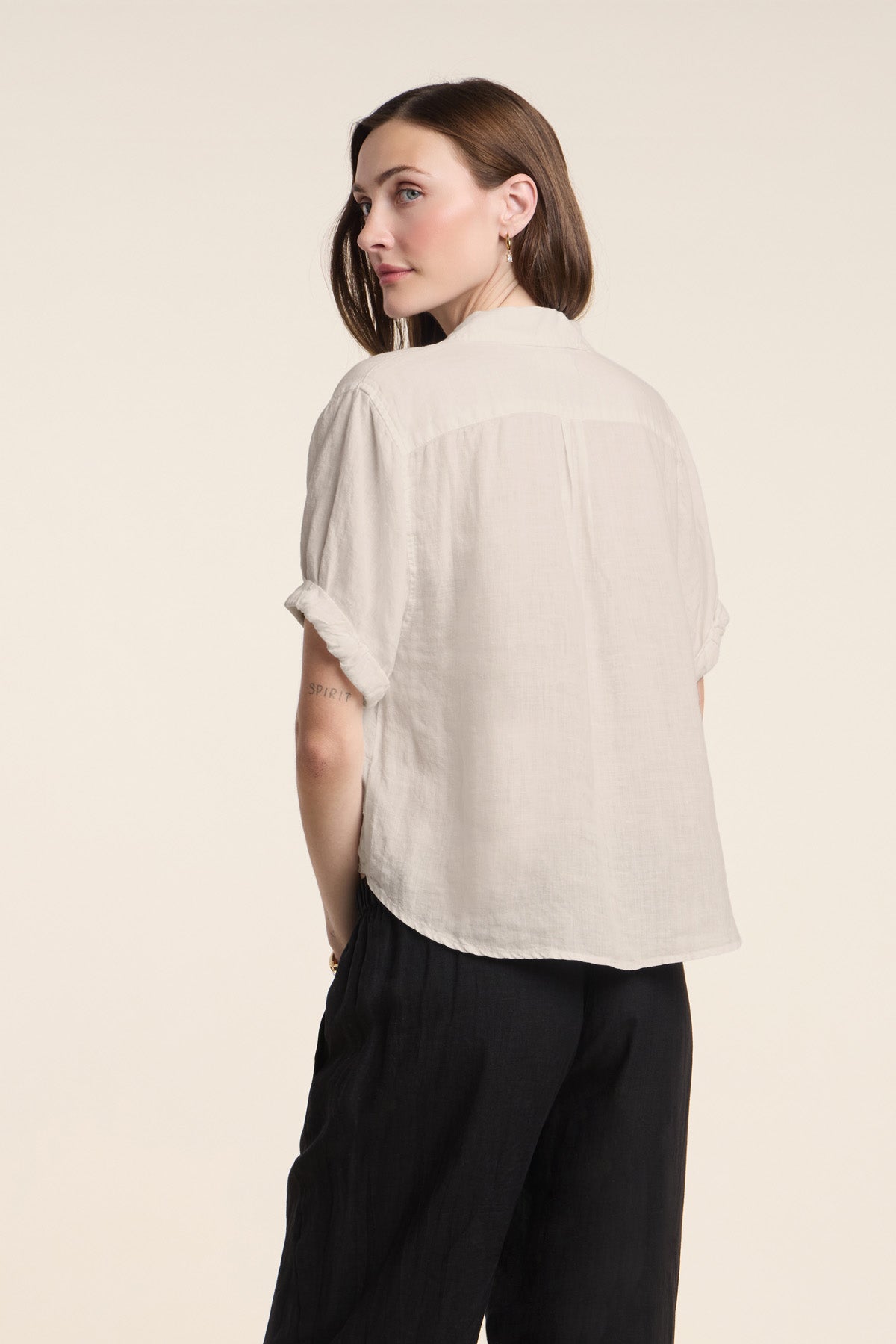 Wearing the VIVICA LINEN TOP by Velvet by Graham & Spencer, a woman with brown hair pairs the lightweight split-neck blouse with black pants as she stands with her back turned and glances over her shoulder—a perfect look for effortless warm-weather dressing.-42119208370369