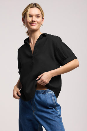 Wearing the VIVICA LINEN TOP by Velvet by Graham & Spencer paired with blue jeans, this individual stands smiling against a plain background, effortlessly showcasing warm-weather style with a relaxed charm.