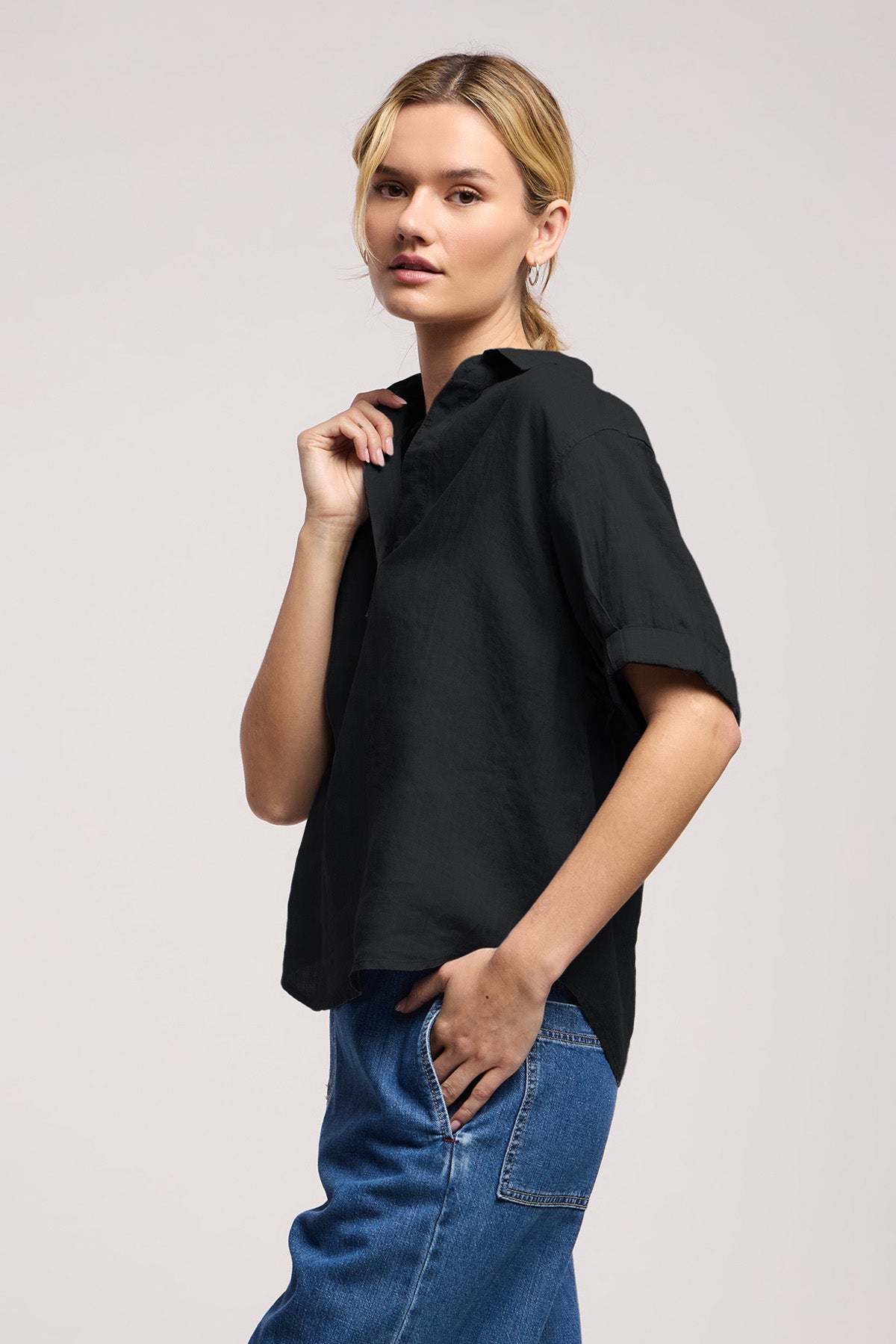 A woman stands slightly sideways against a plain light background, wearing the VIVICA LINEN TOP by Velvet by Graham & Spencer—a lightweight linen piece with a split neckline—paired effortlessly with blue jeans, perfect for warm-weather style.