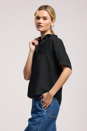 A woman stands slightly sideways against a plain light background, wearing the VIVICA LINEN TOP by Velvet by Graham & Spencer—a lightweight linen piece with a split neckline—paired effortlessly with blue jeans, perfect for warm-weather style.