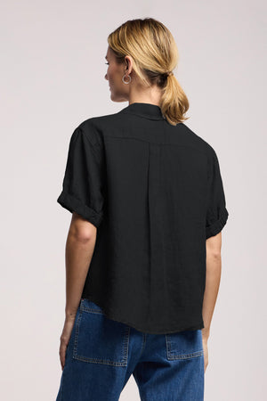 A person with blonde hair in a low ponytail is wearing the VIVICA LINEN TOP by Velvet by Graham & Spencer—a short-sleeve black shirt with a split neckline—paired with blue jeans, facing away from the camera against a plain light background.