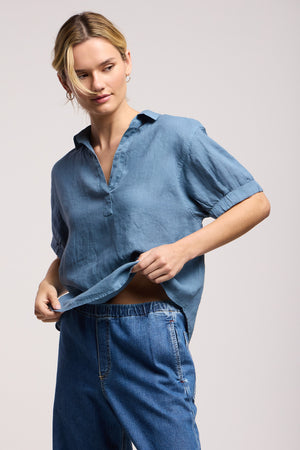 A woman wears the VIVICA LINEN TOP by Velvet by Graham & Spencer—a blue lightweight linen split-neckline shirt—paired with blue jeans against a plain background, perfect for warm-weather style.