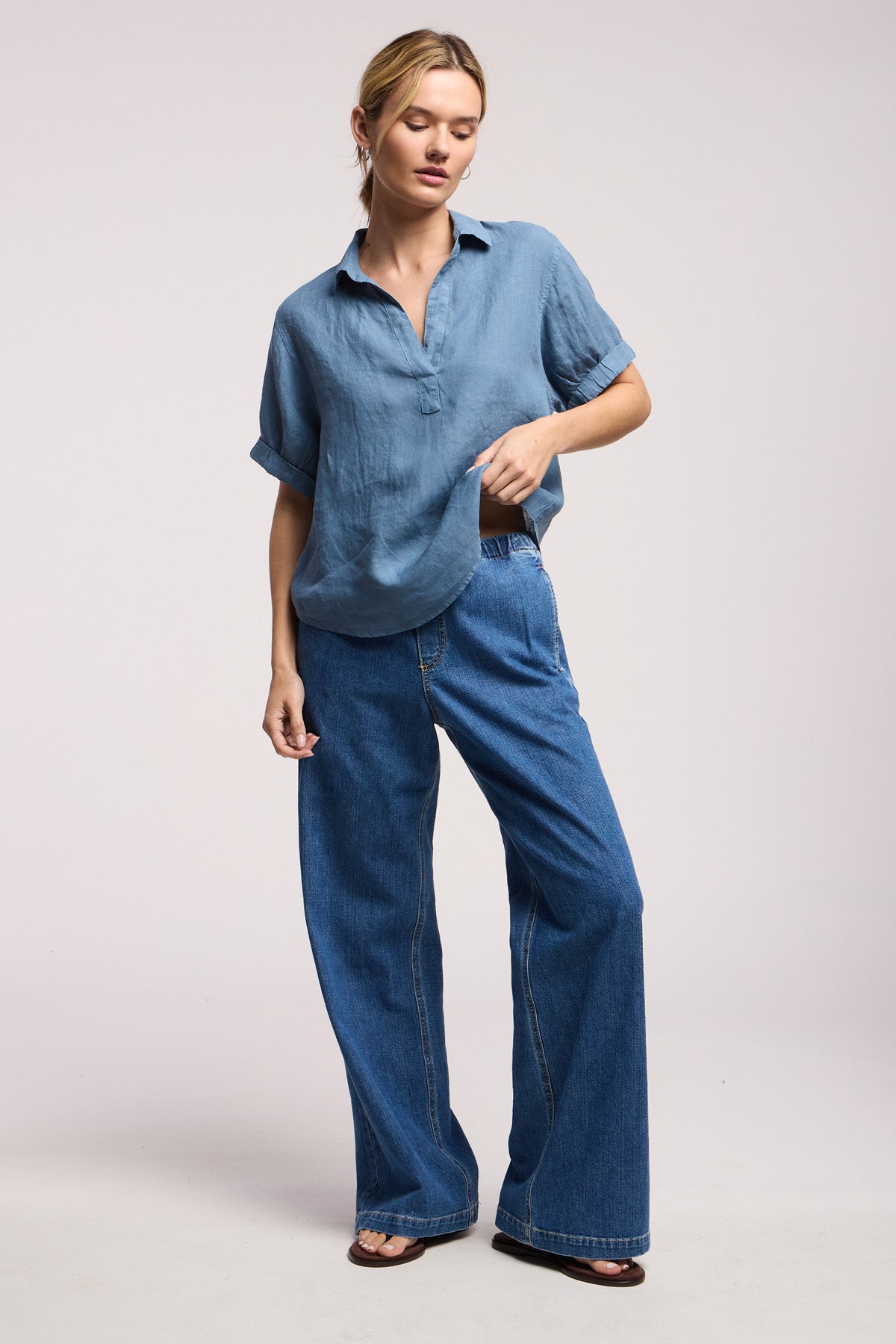 A person stands against a plain background wearing a relaxed fit blue short-sleeve shirt, Velvet by Graham & Spencer SYD JEAN wide-leg denim jeans with an elastic drawstring waist, and brown sandals.