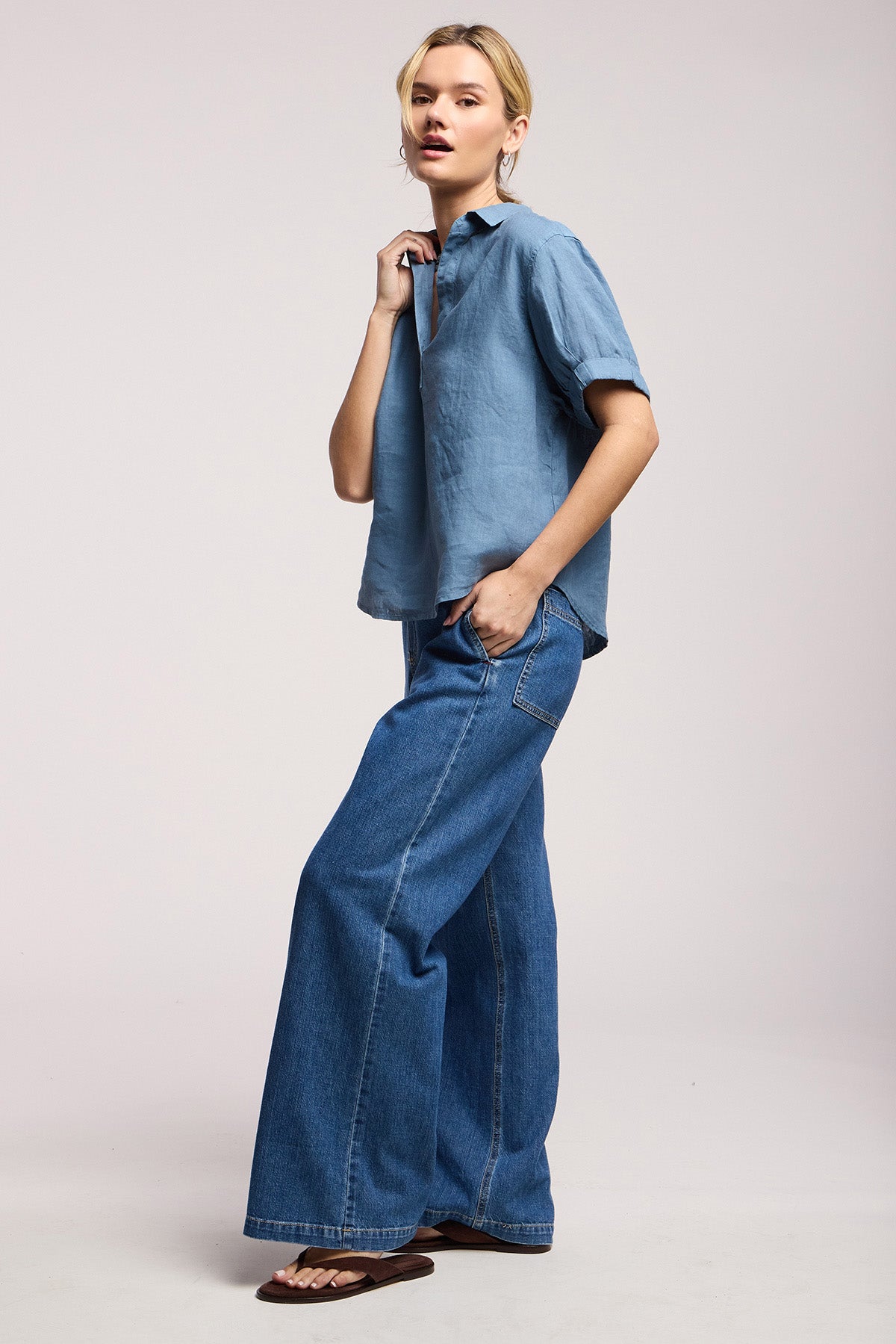 A person stands in profile wearing a light blue short-sleeve shirt, Velvet by Graham & Spencer's SYD JEAN—relaxed fit, wide-leg denim with an elastic drawstring waist—and brown sandals against a plain background.-41527496999105