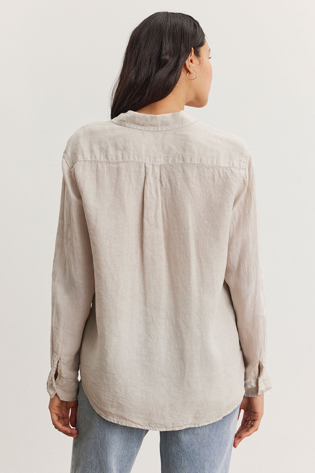 Woman standing facing away, wearing a Velvet by Graham & Spencer Willow Linen Button-Up Shirt and blue jeans, with her long dark hair draped over one shoulder.-36998739132609