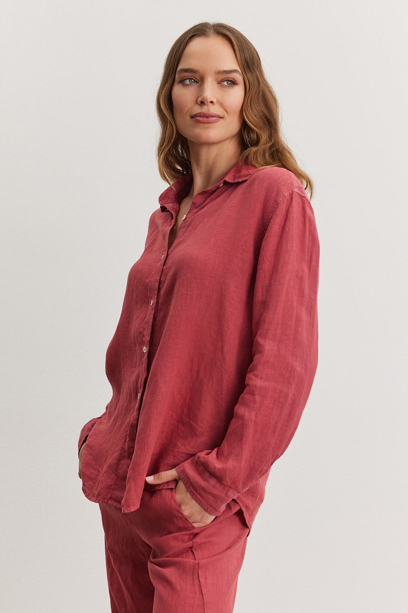 Woman in a relaxed fit red Velvet by Graham & Spencer Willow Linen Button-Up Shirt and pants, standing with one hand in pocket, looking at the camera with a slight smile.