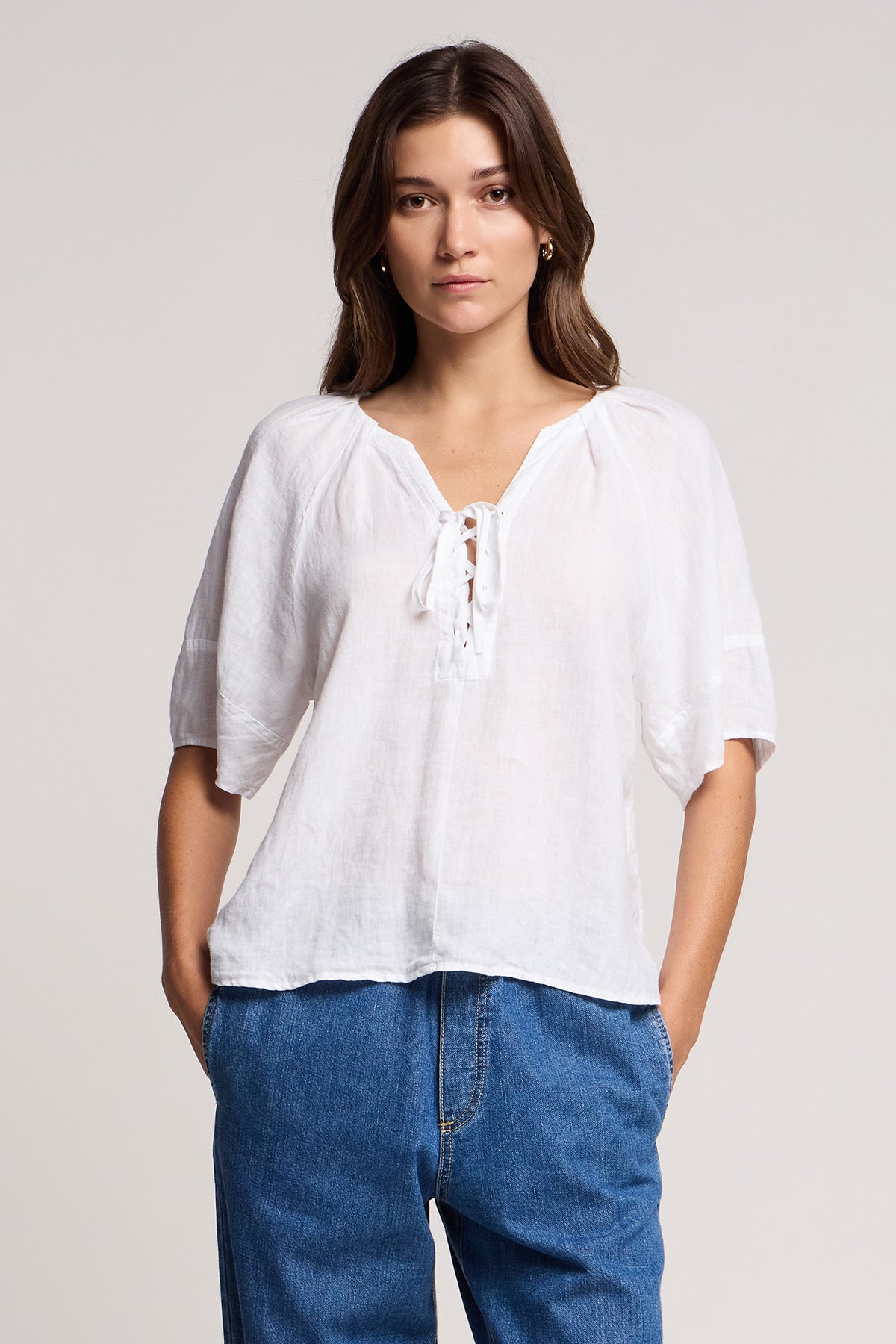 A woman faces forward wearing the FAERYN LINEN TOP by Velvet by Graham & Spencer, a relaxed fit linen design with a lace-up neckline and short sleeves, paired with blue jeans against a plain light background.-41527132946625