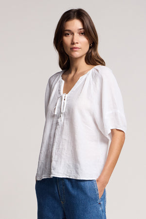 A woman stands with one hand in her pocket, wearing the FAERYN LINEN TOP by Velvet by Graham & Spencer—a relaxed fit linen top with a lace-up neckline—paired with blue jeans against a plain background.