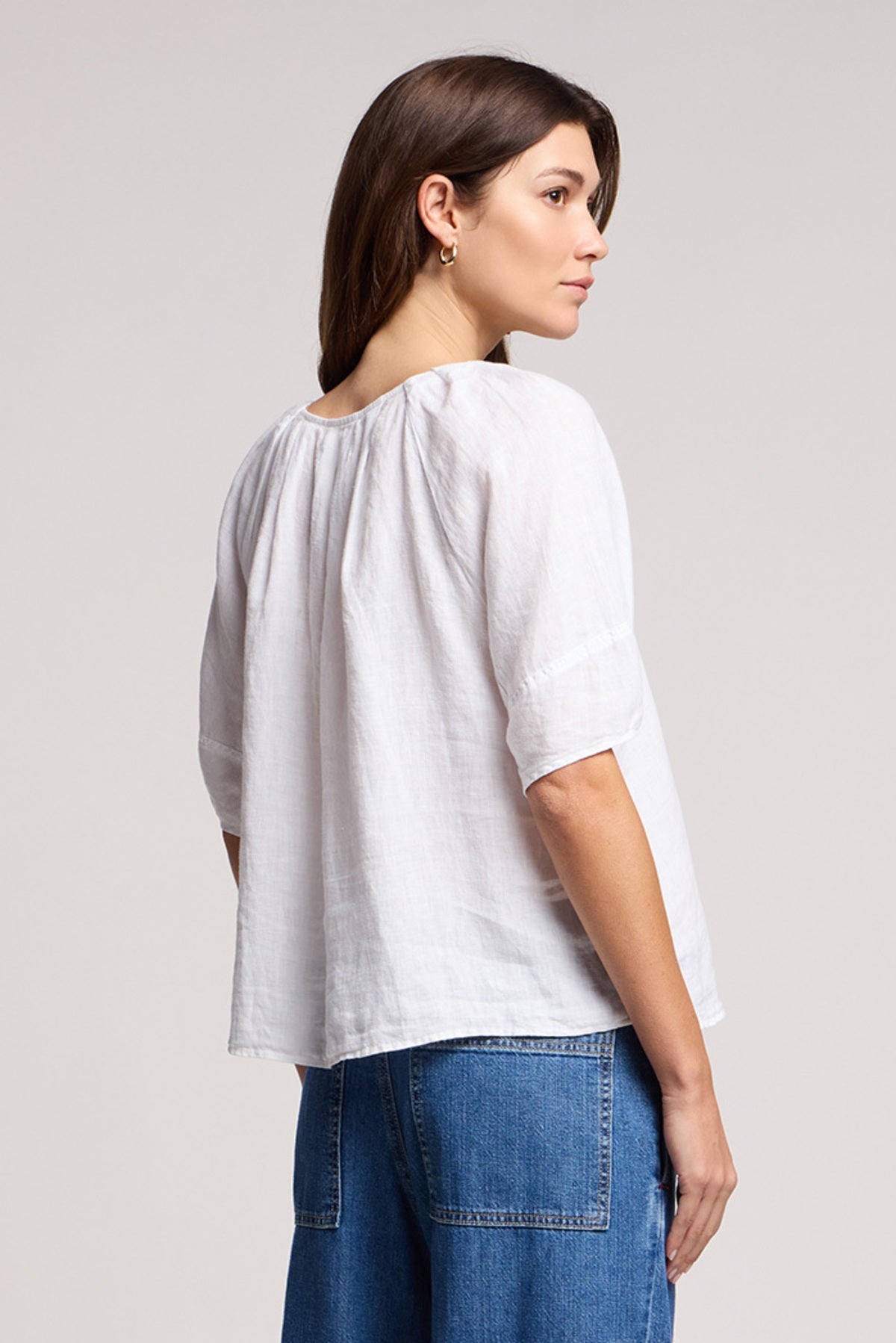 A woman stands sideways wearing the FAERYN LINEN TOP by Velvet by Graham & Spencer—featuring a relaxed fit and elbow-length sleeves—paired with blue jeans against a plain background.