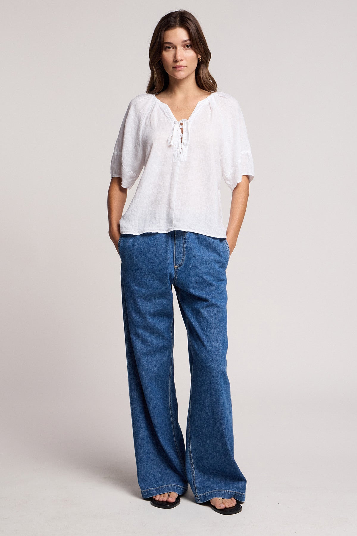 A woman stands facing forward, wearing Velvet by Graham & Spencer's SYD JEAN—relaxed fit, wide-leg denim with an elastic drawstring waist—paired with a white short-sleeve tie-neck blouse and black sandals against a light background.
