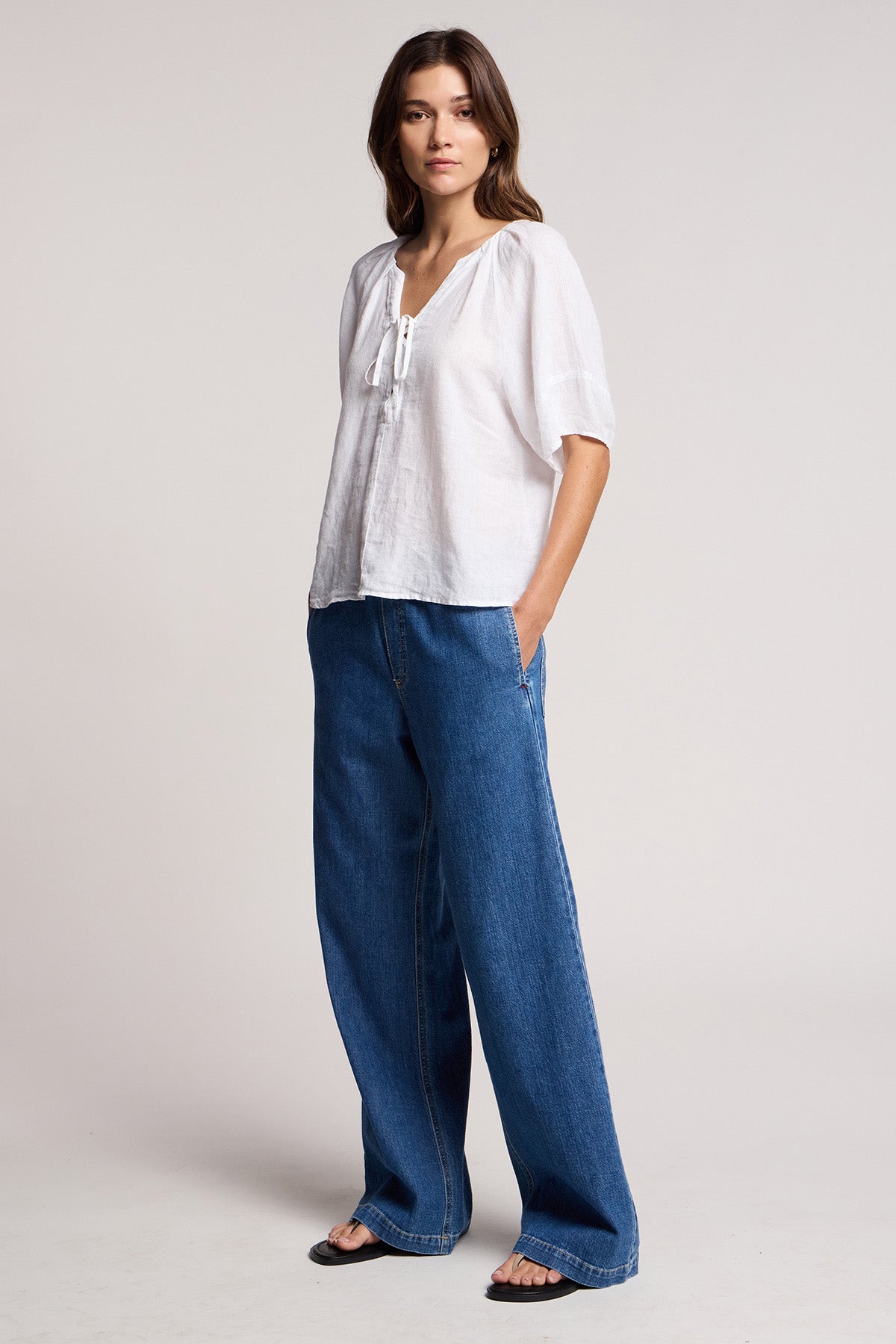 A person stands against a plain background wearing a loose white blouse, Velvet by Graham & Spencer’s SYD JEAN—relaxed fit wide-leg denim jeans with an elastic drawstring waist—and black sandals.-41527496900801