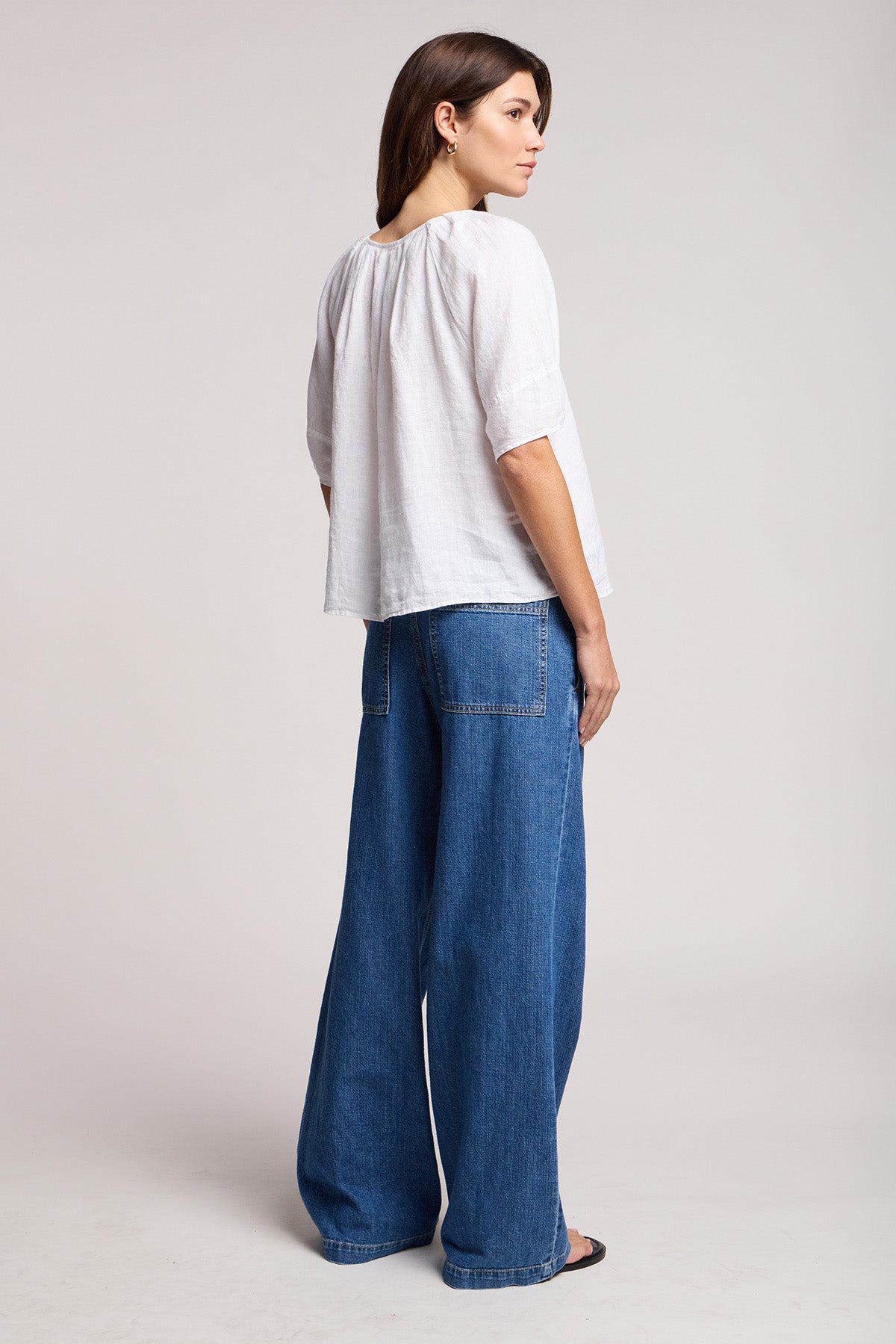 A woman stands sideways in the SYD JEAN by Velvet by Graham & Spencer, featuring relaxed-fit wide-leg denim with an elastic drawstring waist, visible back pockets, and a loose white top against a plain light background.-41527496933569