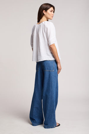 A woman stands sideways in the SYD JEAN by Velvet by Graham & Spencer, featuring relaxed-fit wide-leg denim with an elastic drawstring waist, visible back pockets, and a loose white top against a plain light background.