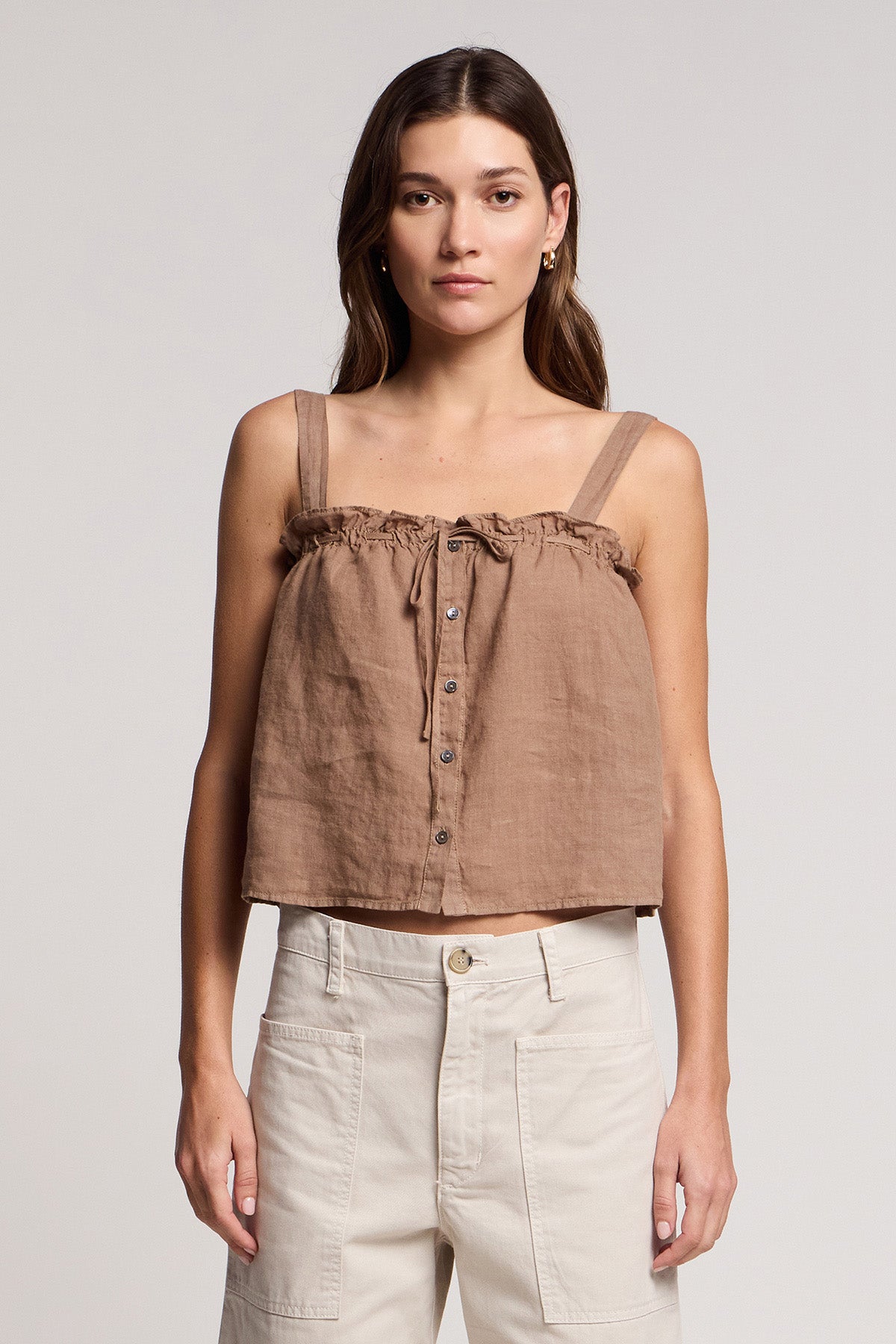 A woman wears the IZZIE LINEN TANK TOP by Velvet by Graham & Spencer—a sleeveless, brown, button-up crop with ruffle details—paired with high-waisted, light pants against a plain background.-41527050272961