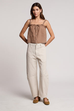 A woman stands against a plain background wearing the IZZIE LINEN TANK TOP by Velvet by Graham & Spencer, styled with light high-waisted pants and brown flat shoes.