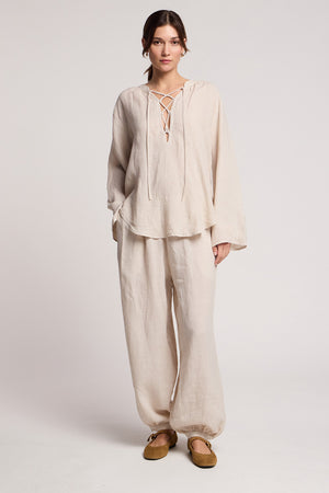 A woman stands in a studio wearing Velvet by Graham & Spencer's CAMBER LINEN PANT with an elastic waist, paired with a loose-fitting woven linen top and brown flats.