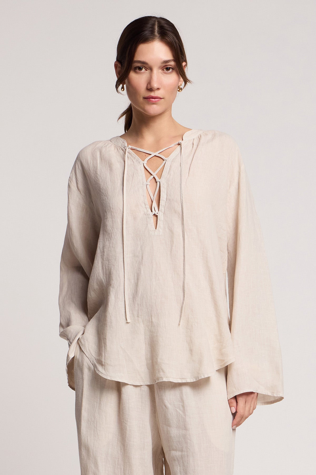 A woman stands against a plain background wearing Velvet by Graham & Spencer’s cream CAMBER LINEN PANT with an elastic waist, paired with a loose, long-sleeve linen top featuring a lace-up neckline.