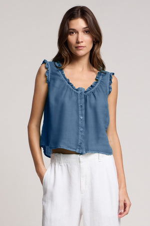 A woman is wearing the TANA LINEN TOP by Velvet by Graham & Spencer—a sleeveless blue ruffle trim top with button-up detail—paired with white high-waisted pants, standing against a plain background.