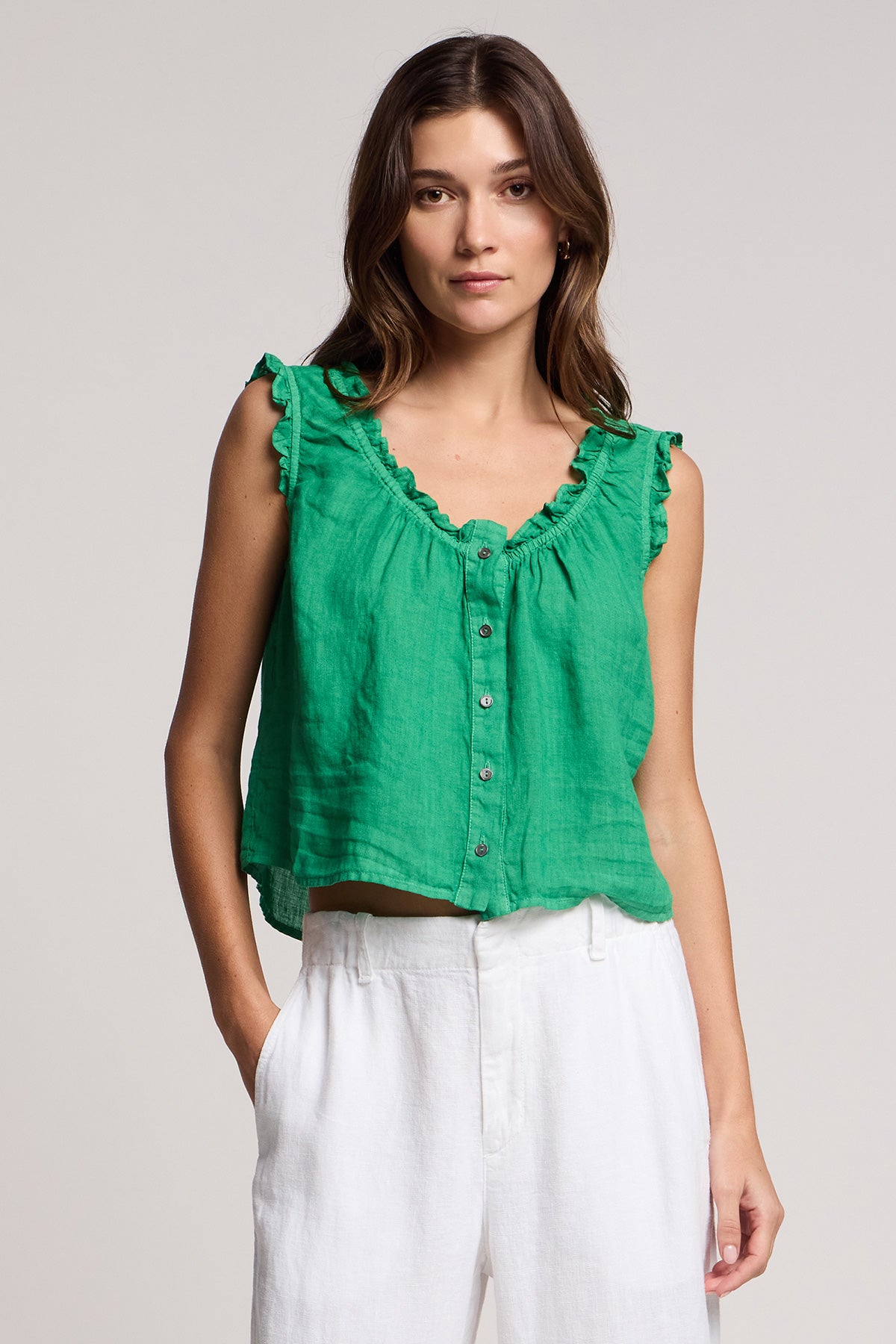 A woman wearing the TANA LINEN TOP by Velvet by Graham & Spencer, a sleeveless green ruffled button-up, paired with white pants against a plain light background.-41527238262977
