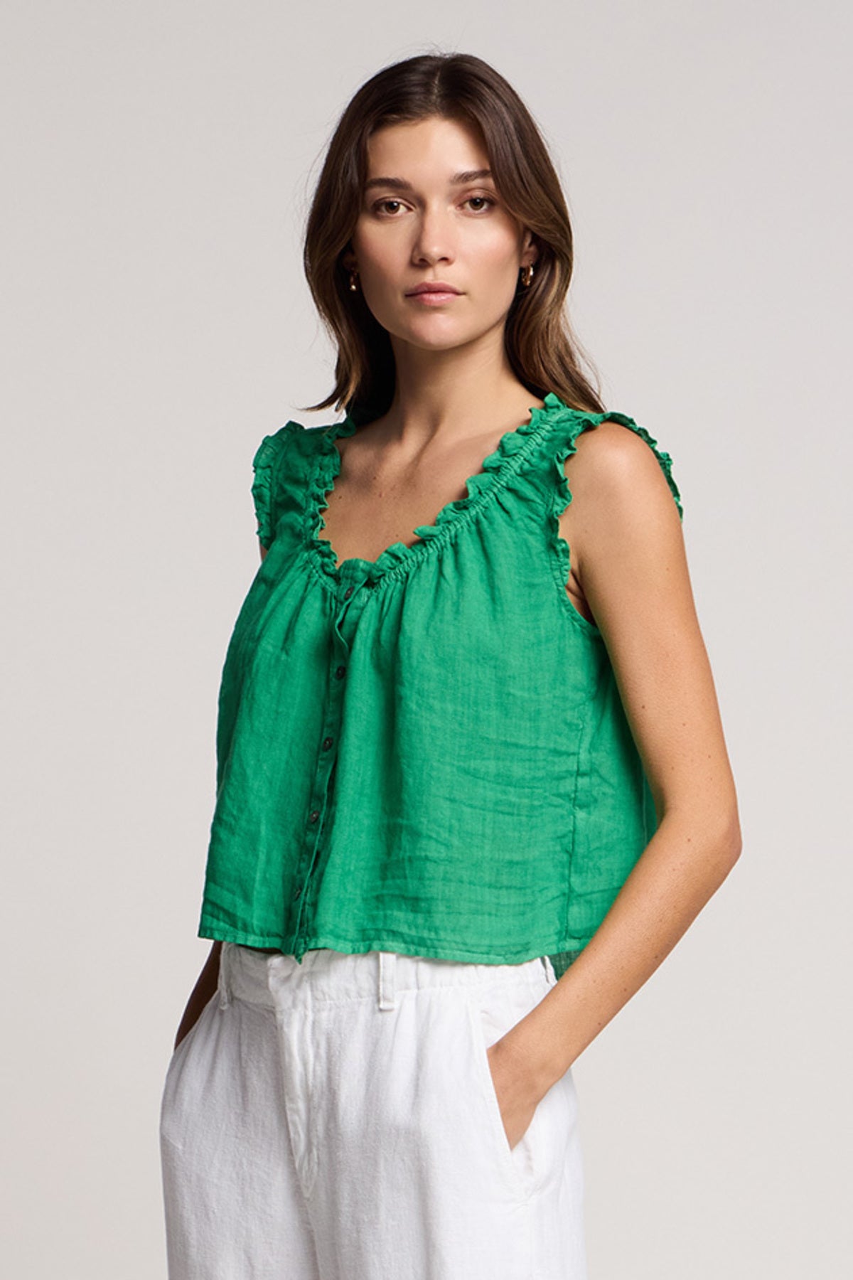 Woman wears the TANA LINEN TOP by Velvet by Graham & Spencer, a green sleeveless ruffled blouse, paired with white pants and standing with hands in pockets against a plain background.