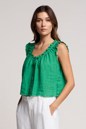 Woman wears the TANA LINEN TOP by Velvet by Graham & Spencer, a green sleeveless ruffled blouse, paired with white pants and standing with hands in pockets against a plain background.