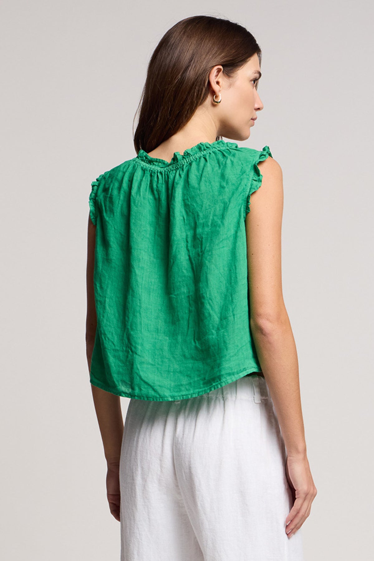 A woman with straight brown hair is seen from the back, wearing Velvet by Graham & Spencer's TANA LINEN TOP—a sleeveless green top with ruffle details—paired with loose white pants against a plain background.-41527238295745