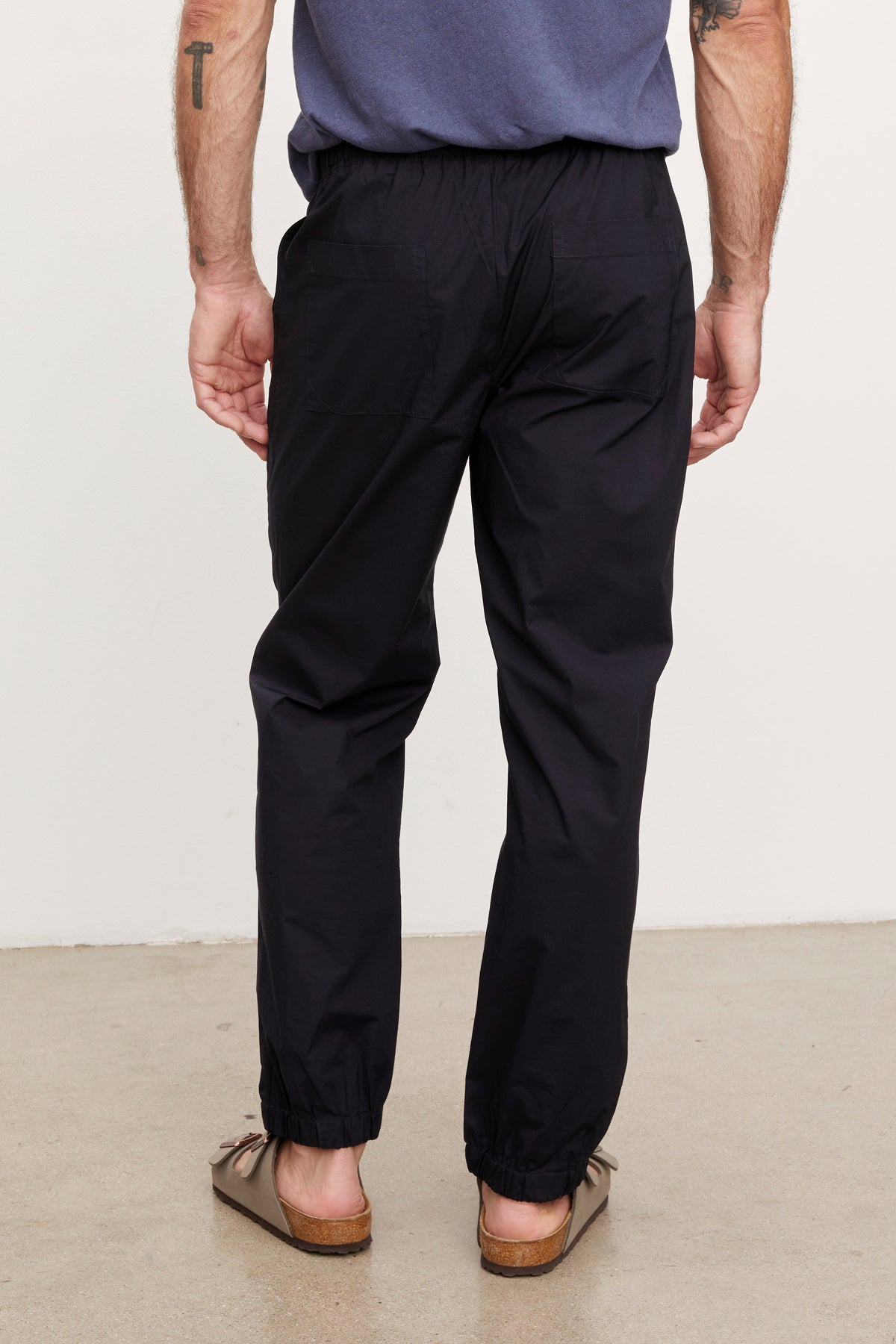 Man standing, viewed from the back, wearing black Velvet by Graham & Spencer Lazarus Jogger ankle trousers and brown shoes, hands slightly behind him.-36732528885953