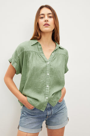 The model is wearing a Velvet by Graham & Spencer ARIA LINEN BUTTON FRONT TOP in sage green and denim shorts, exuding timeless appeal.