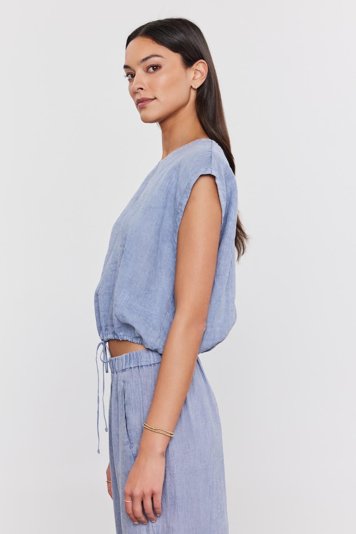   A woman in a Velvet by Graham & Spencer BAYLA LINEN TOP with a scoop neckline and matching pants, looking away from the camera with her hair in a low ponytail. 