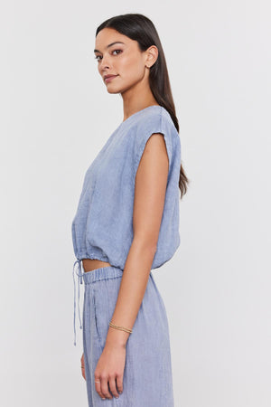 A woman in a Velvet by Graham & Spencer BAYLA LINEN TOP with a scoop neckline and matching pants, looking away from the camera with her hair in a low ponytail.