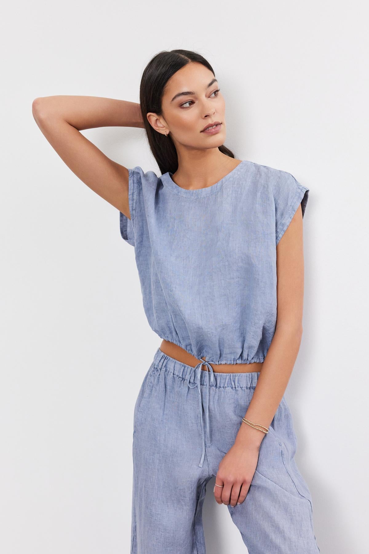   A woman in a stylish blue denim jumpsuit with a cocoon silhouette, posing with one hand raised to her head against a plain white background by Velvet by Graham & Spencer. 