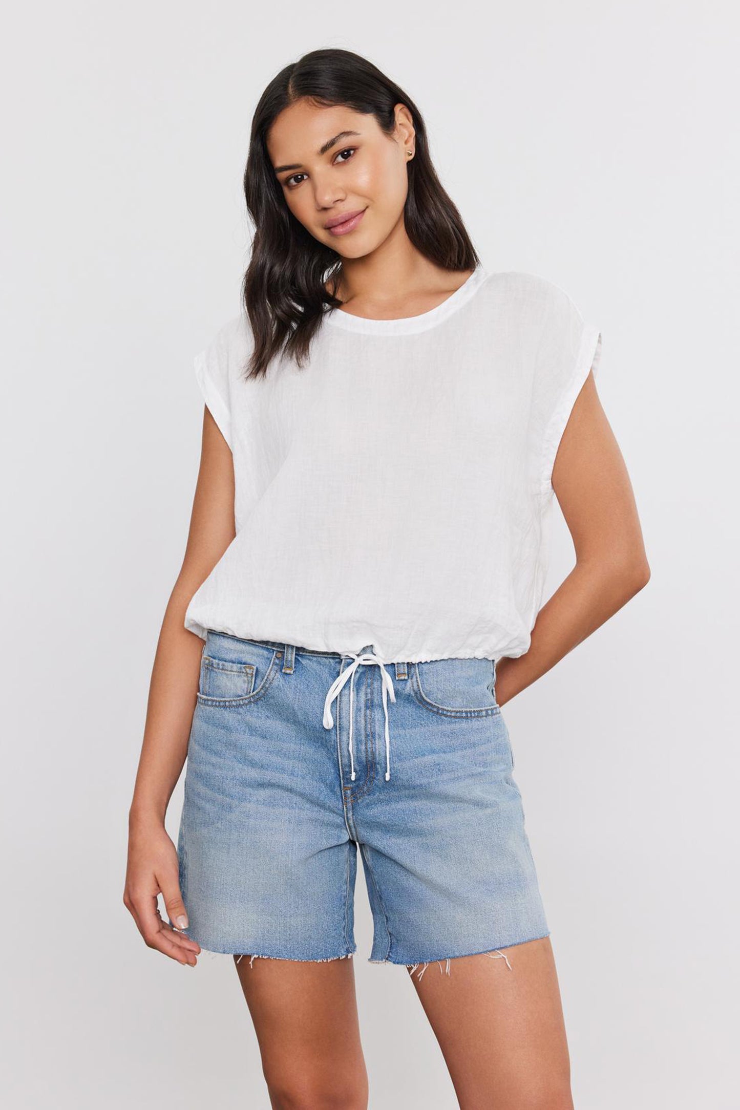 A woman wearing a Velvet by Graham & Spencer's BAYLA LINEN TOP with a scoop neckline and denim shorts, standing against a plain background, looking at the camera with a slight smile.-36752900751553