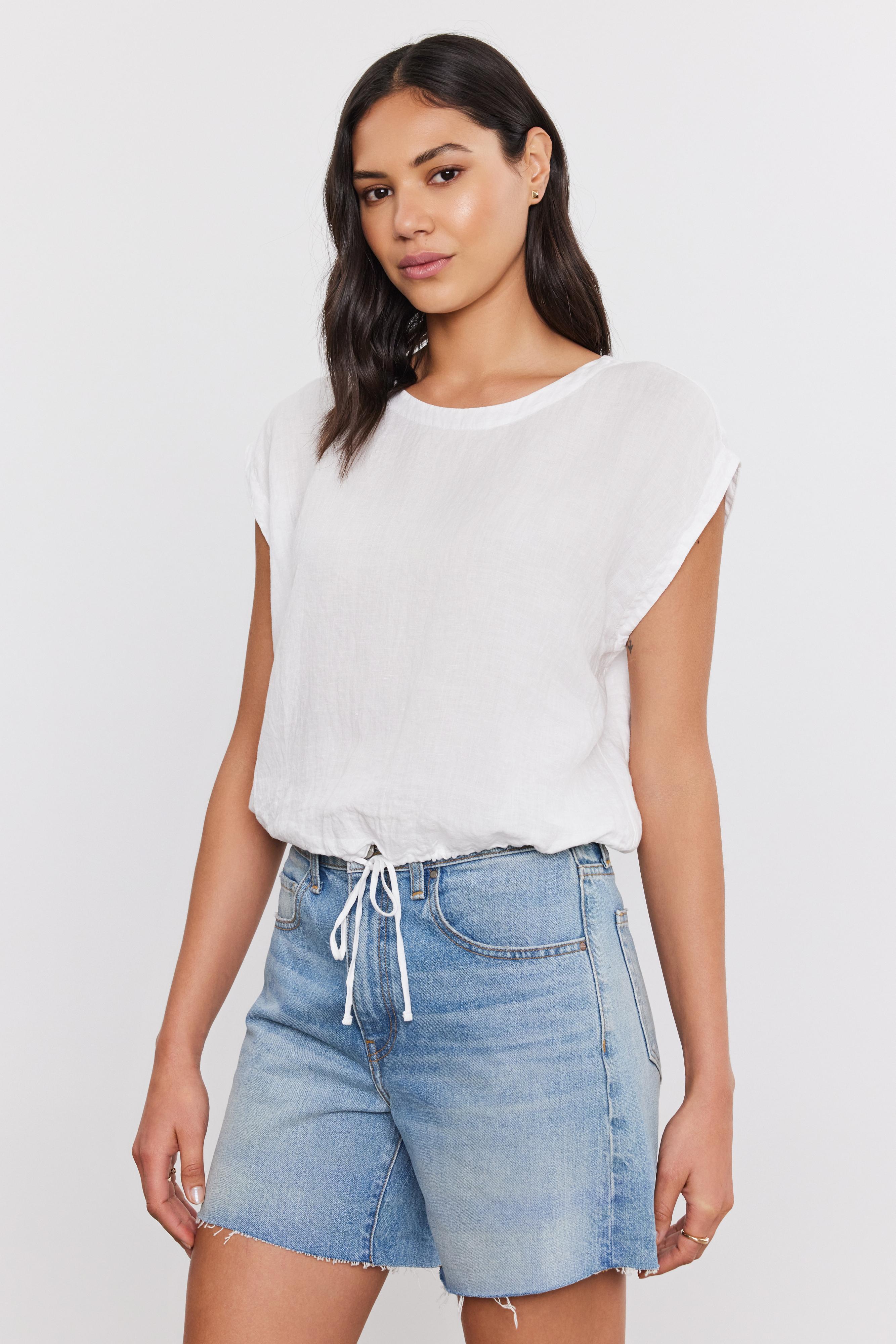   Woman wearing a BAYLA LINEN TOP from Velvet by Graham & Spencer with a scoop neckline and blue denim shorts standing against a plain background. 