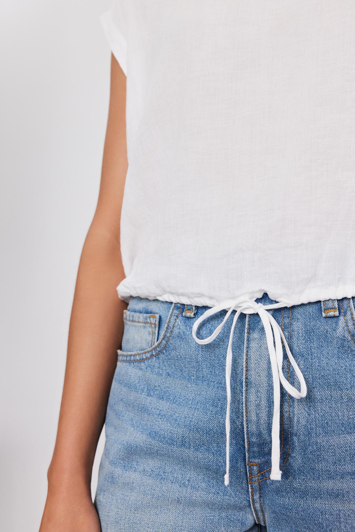   Close-up of a person wearing a Velvet by Graham & Spencer BAYLA LINEN TOP and blue denim jeans with a drawstring cinch. 