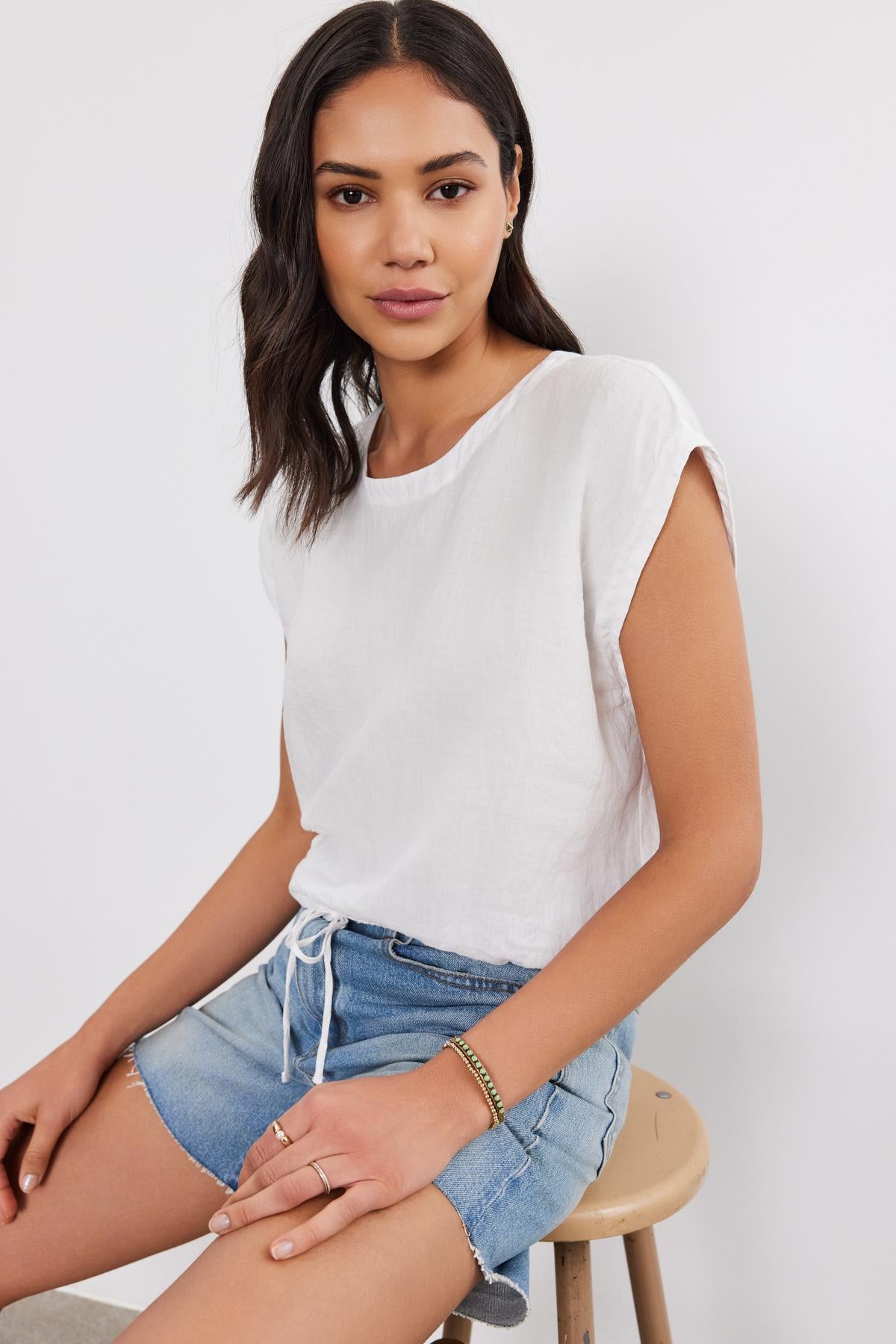   A woman in a Velvet by Graham & Spencer BAYLA LINEN TOP and denim shorts sitting on a stool, looking at the camera with a slight smile. 