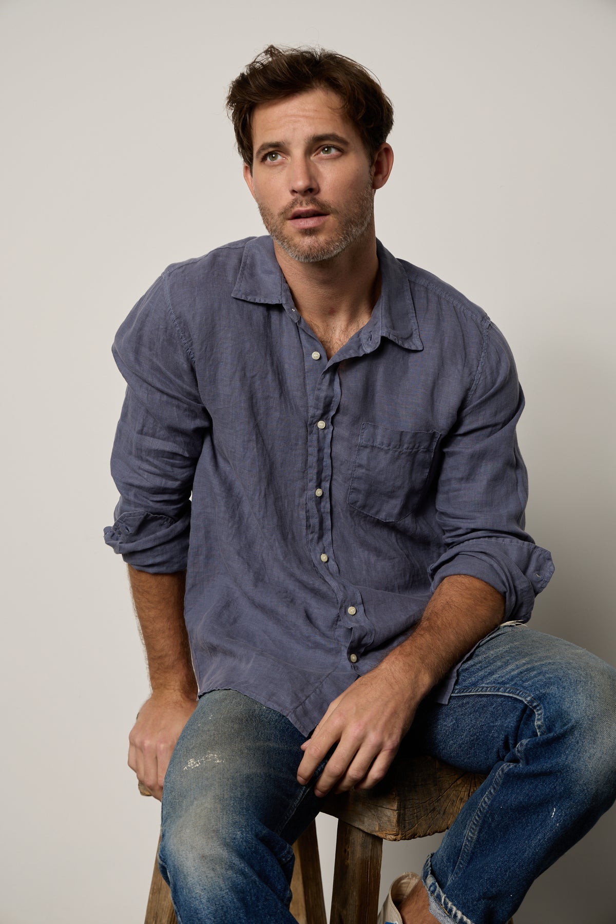 A man in jeans and a BENTON LINEN BUTTON-UP SHIRT by Velvet by Graham & Spencer sitting on a stool.-35955069354177