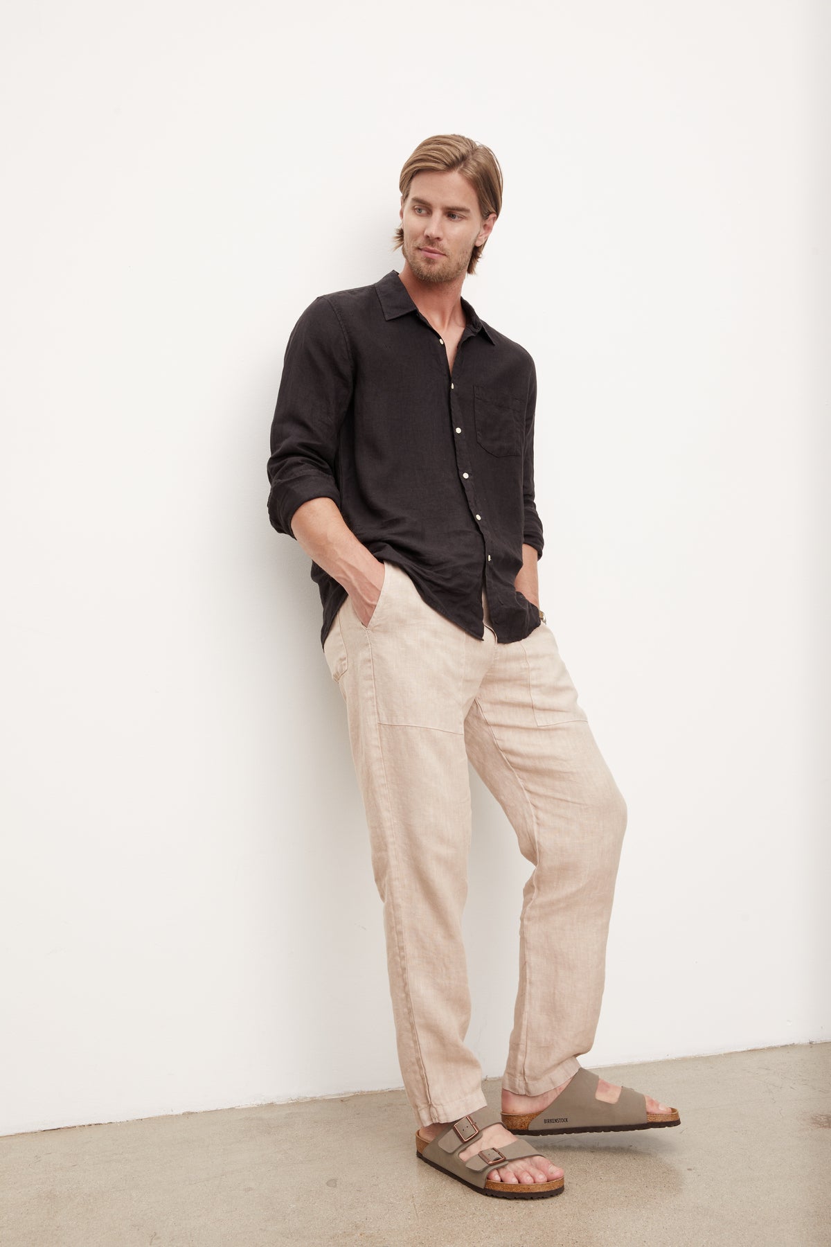 A stylish man leaning against a wall in a Velvet by Graham & Spencer BENTON LINEN BUTTON-UP SHIRT.-36009360556225