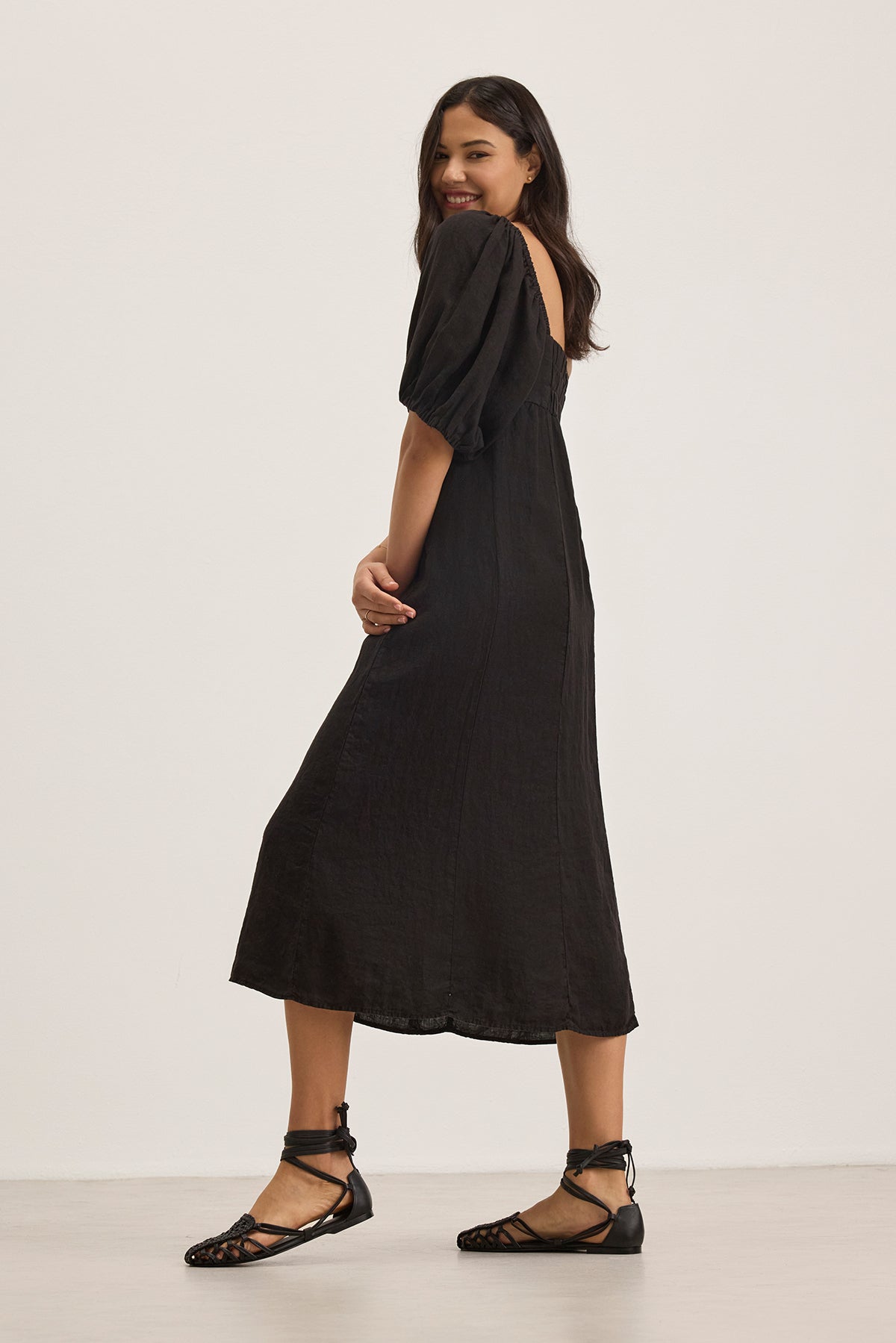 A woman in the BROOKE LINEN DRESS by Velvet by Graham & Spencer with puff sleeves and lace-up sandals smiles, standing against a plain light background.-38662646792385
