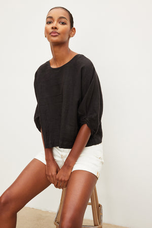 A woman is sitting on a stool wearing a Velvet by Graham & Spencer CASS LINEN CREW NECK TOP with a twist knot and white shorts.